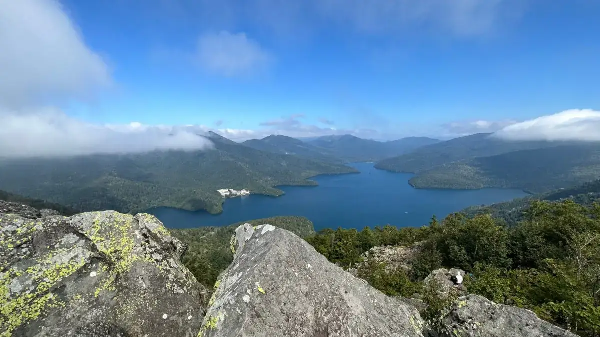 Photo of A panoramic view of a mountain lake surrounded by hills and forests under a clear blue sky. | Wander Sober